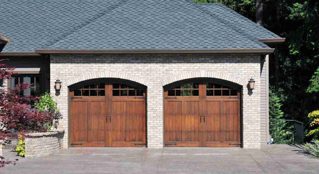 Two custom wooden carriage-house style garage doors with windows and decorative hardware installed on a brick home, showcasing stylish and durable garage door upgrades.