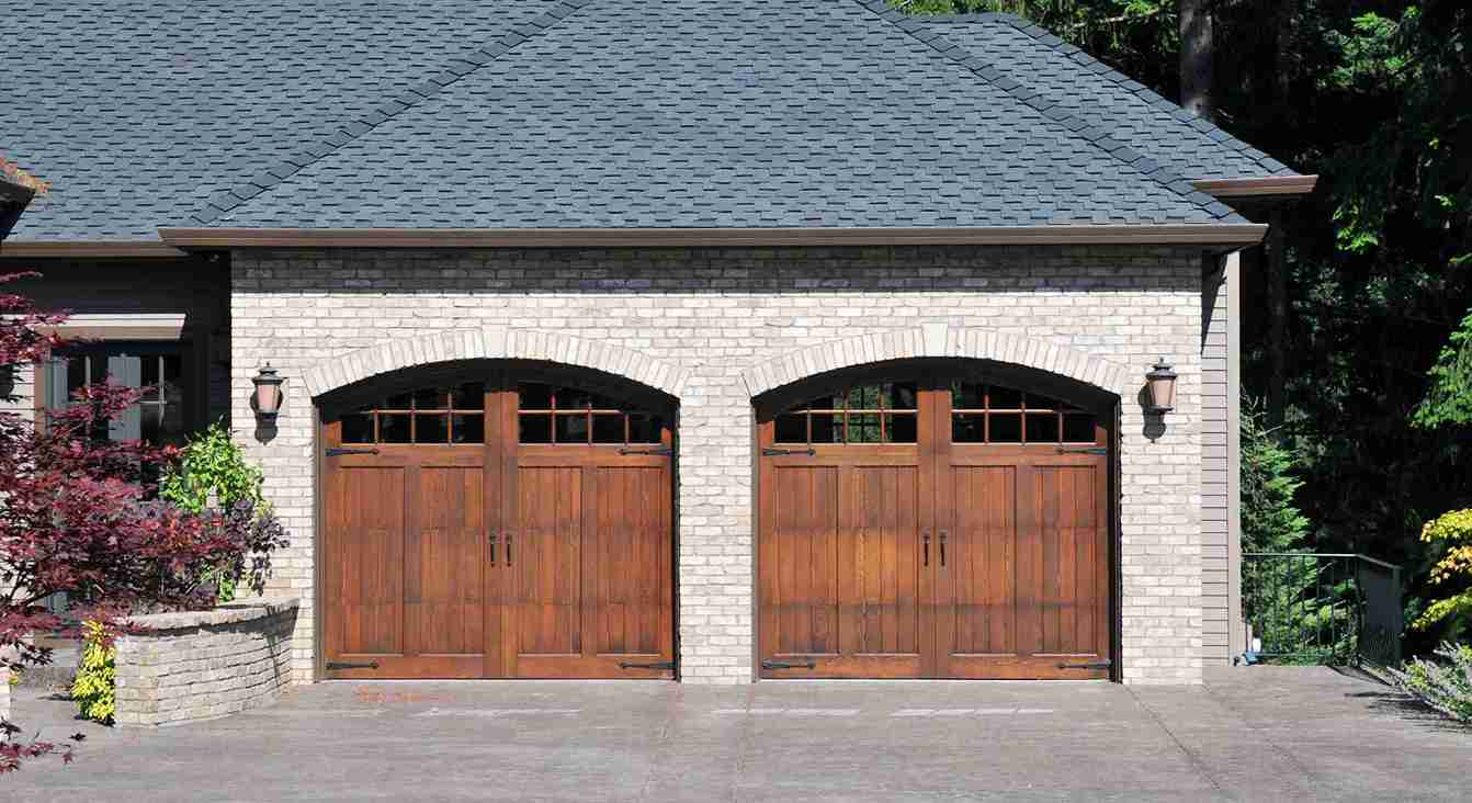 Two custom wooden carriage-house style garage doors with windows and decorative hardware installed on a brick home, showcasing stylish and durable garage door upgrades.